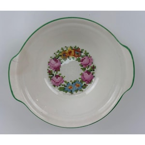 May include: A white ceramic bowl with a green rim and a floral design in the center. The design is a wreath of pink flowers with green leaves and a yellow center.