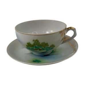 May include: White teacup and saucer set with a painted landscape scene. The cup and saucer feature a gold trim and a handle with gold accents. The landscape includes green trees and blue water.