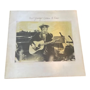 Puede incluir: Portada del álbum "Comes a Time" de Neil Young. La imagen es una fotografía en blanco y negro de Neil Young tocando una guitarra acústica en el escenario. La portada del álbum tiene un borde blanco y el título está escrito en una fuente manuscrita.