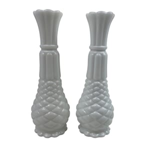 Puede incluir: Un par de jarrones de vidrio lechoso blanco a juego con un patrón de diamante texturizado similar a una piña en la parte inferior, un cuello liso con nervaduras verticales y un borde festoneado. Estos recipientes altos y delgados tienen un acabado brillante.