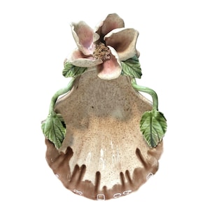Könnte beinhalten: Eine muschelförmige Keramikschale mit einer großen, detaillierten Blume und grünen Blättern. Die Blume hat rosa und cremefarbene Blütenblätter, und die Schale hat eine gesprenkelte, braune und cremefarbene Oberfläche. Es ist ein dekorativer Artikel.