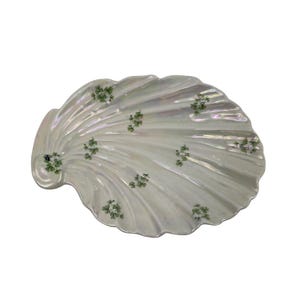 Könnte beinhalten: Eine muschelförmige Schale mit irisierender, perlmutteigener Oberfläche. Das Design zeigt ein wiederkehrendes Muster aus kleinen, grünen dreiblättrigen Kleeblattmotiven, die über die Rillen und Erhöhungen der Keramik verteilt sind.