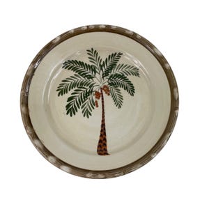 May include: A ceramic plate with a cream-colored center featuring a hand-painted palm tree design. The palm tree has green fronds, a brown trunk, and orange fruit. The rim of the plate has a brown bamboo-like design.