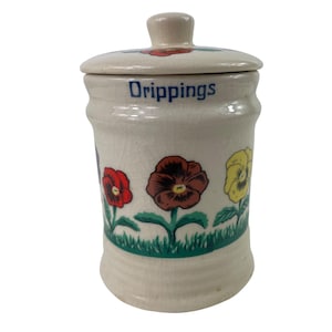 May include: Cream-colored ceramic jar with lid, featuring the word "Drippings" in blue. The jar is decorated with red, brown, and yellow pansies above a green grass border. The lid has a small knob and a red flower.