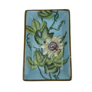 Puede incluir: Una bandeja rectangular con fondo azul claro que presenta una ilustración detallada de una flor de la pasión con hojas verdes y zarcillos. La bandeja está enmarcada por un borde metálico fino de tono dorado que perfila el borde.