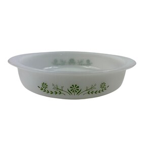 May include: A round, white glass baking dish with a shallow profile and integrated side handles. The exterior features a repeating green botanical pattern consisting of stylised flowers and delicate leafy sprigs centred on the side wall.