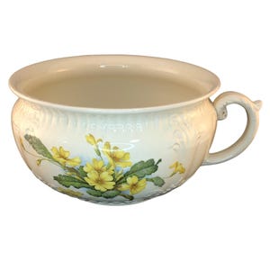 May include: Cream-colored ceramic bowl with a handle, decorated with yellow primrose flowers and green leaves. The bowl has a wide opening and a decorative embossed pattern around the rim. A classic piece for serving or display.