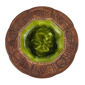 May include: A brown ceramic ashtray with a green glazed center. The ashtray is decorated with images of San Francisco landmarks, including Alcatraz, the Golden Gate Bridge, Chinatown, and Lombard Street.