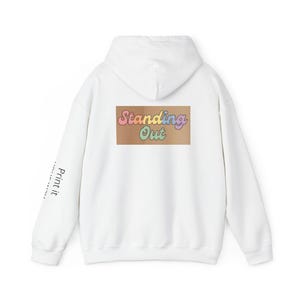 Op de afbeelding: Witte hoodie met een capuchon en lange mouwen. De achterkant heeft een rechthoekig ontwerp met de woorden "Standing Out" in pastelkleuren. De linkermouw heeft verticaal de tekst "Print it".