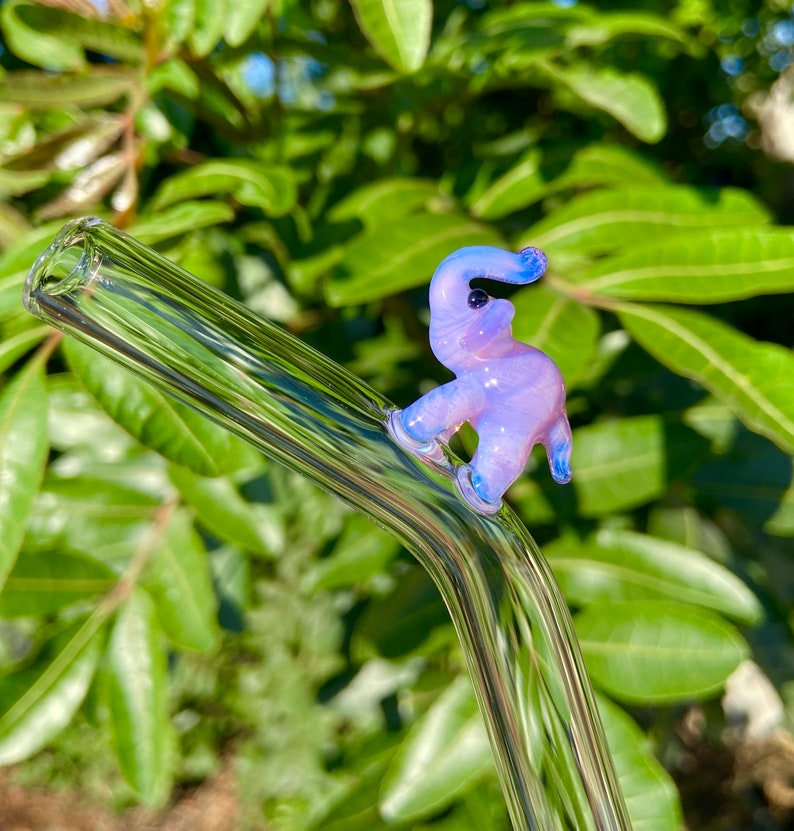 Pu&ograve; includere: Una cannuccia da bere in vetro trasparente con un ciondolo a forma di elefante in vetro viola e blu attaccato alla parte superiore.