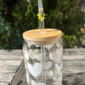 Puede incluir: Un vaso de vidrio transparente con tapa de bambú y pajita de vidrio. El vaso está decorado con diseños de corazones blancos. La pajita tiene un adorno de girasol amarillo y negro. El vaso está sobre una superficie de madera.
