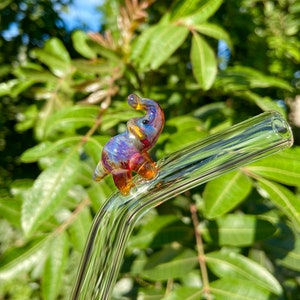 Pu&ograve; includere: Una cannuccia da bere in vetro trasparente con una statuetta di elefante in vetro colorato fissata in cima. L'elefante &egrave; iridescente con sfumature di blu, viola e arancione.