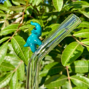 Pu&ograve; includere: Una statuetta di elefante in vetro blu turchese appoggiata su una cannuccia da bere in vetro trasparente.