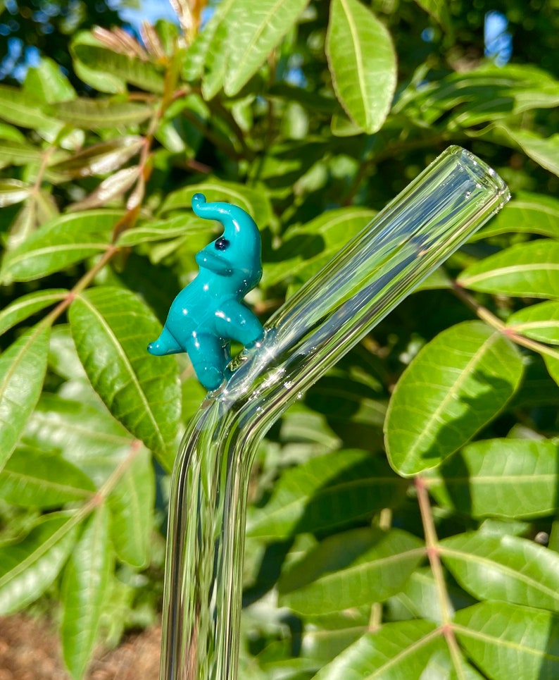Pu&ograve; includere: Una statuetta di elefante in vetro blu turchese appoggiata su una cannuccia da bere in vetro trasparente.