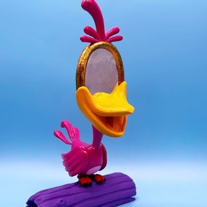 May include: A pink and yellow bird figurine with a gold mirror on its head, perched on a purple log. The bird has a wide, open beak and a cheerful expression.