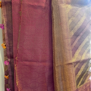 May include: A draped textile featuring a deep magenta hue with intricate circular patterns and gold accents. The fabric has a sheen, with a contrasting gold and brown border. Colorful tassels adorn the edge, adding a decorative touch.
