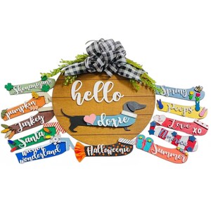 May include: A wooden round door hanger with the text "hello doxie" and a dachshund wearing a blue sweater with a pink heart.  The door hanger has interchangeable seasonal greetings, including "spring", "peeps", "love", "america", "summer", "halloweenie", "wonderland", "santa", "turkey", "pumpkin", and "shenanigans".