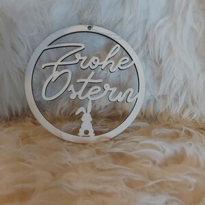 May include: White wooden circle ornament with the text "Frohe Ostern" and a silhouette of a bunny rabbit.