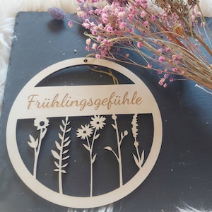 May include: A round wooden sign with the text "Frühlingsgefühle" and a design of five stylized flowers. The sign is hanging on a black background.