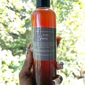 May include: A brown glass bottle with a black cap and label. The label reads "MADE WITH ESSENTIAL OILS PIPE Cleaner". The bottle is filled with a dark red liquid.