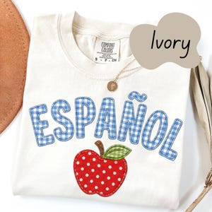 May include: An ivory t-shirt featuring the word ESPAÑOL in blue and white gingham letters across the chest. Below the text is an appliqué of a red apple with white polka dots and a green gingham leaf. The shirt has a Comfort Colors brand label.