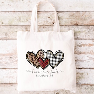 May include: Off-white tote bag with three heart designs. The hearts feature leopard print, red plaid, and black and white buffalo check patterns. The text "Love never fails" and "1 Corinthians 13:8" are printed below the hearts.