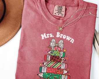 May include: A maroon Comfort Colors t-shirt with the name "Mrs. Brown" printed on it. The shirt features a graphic of a stack of pencils decorated as a Christmas tree with a white bow, lights, and stars. A silver necklace with a pendant is on the shirt.