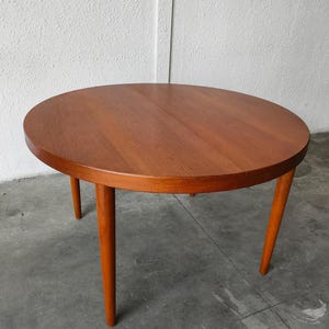 Może przedstawiać: Okrągły drewniany stół w stylu mid-century modern. Stół ma czerwonobrązowe wykończenie i okrągły blat wsparty na czterech zwężanych nogach. Średnica stołu wynosi około 61 cm.