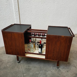 Può includere: Un carrello bar moderno di metà secolo con finitura in legno scuro e superfici superiori nere. Il carrello presenta un interno a specchio, portabicchieri ed è fornito di bottiglie e accessori da bar. È su piccole ruote.