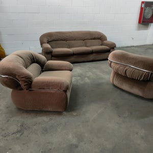May include: A brown three-piece living room set with a sofa and two armchairs. The furniture is upholstered in a soft, textured fabric and has a modern design.