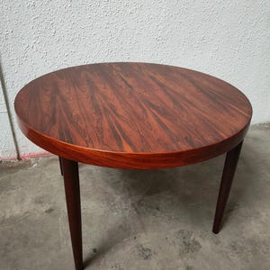 Può includere: Un tavolo rotondo in stile moderno di metà secolo con finitura in legno scuro. Il tavolo ha un diametro di circa 76 cm ed è sostenuto da quattro gambe affusolate e sottili. La venatura del legno presenta una ricca tonalità marrone-rossastra.