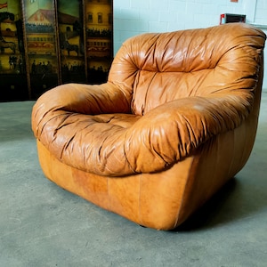 Op de afbeelding: Een grote, afgeronde vintage fauteuil bekleed met verweerd, lichtbruin leer. De stoel heeft een laag profiel met een brede zitting en zwaar gevoerde armleuningen en rugleuning. Het leer heeft een gekreukte textuur.