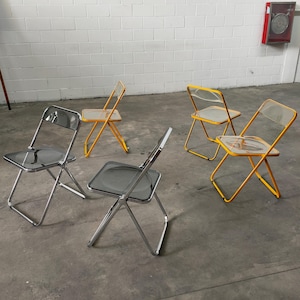 Op de afbeelding: Vier opvouwbare stoelen met doorzichtige kunststof zittingen en metalen frames. Twee stoelen zijn geel en twee stoelen zijn zilver.