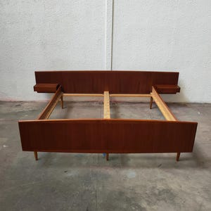 Pode incluir: Uma estrutura de cama de madeira de estilo moderno de meados do século. A cama tem uma cabeceira com mesas de cabeceira integradas. A estrutura é de cor castanha quente e tem um design de baixo perfil. A estrutura da cama é suportada por pernas de madeira. Mede cerca de 200 cm de comprimento.