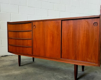 May include: A mid-century modern teak sideboard with three drawers and two doors. The sideboard has a warm brown finish and sits on four legs.