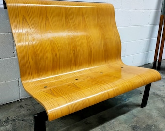 Könnte beinhalten: Eine Holzbank mit einer geschwungenen Rückenlehne und schwarzen Metallbeinen. Die Bank ist aus hellem Holz mit natürlicher Maserung.