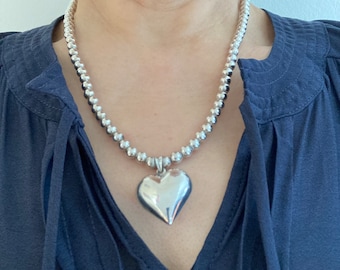 May include: A silver beaded necklace with a large, polished heart pendant. The necklace features small, round beads and a prominent heart-shaped charm, reflecting light. The jewelry is displayed against a dark blue background.