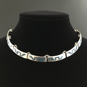 May include: A silver-toned necklace with a geometric design. The necklace is composed of rectangular links connected by small rings. Each link features a turquoise-colored inlay in a zig-zag pattern. The necklace is displayed on a black mannequin.