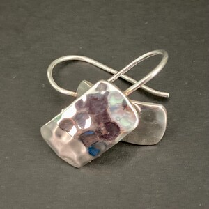 May include: A pair of silver earrings featuring rectangular, hammered metal drops with a reflective, uneven surface. Each earring is attached to a curved, thin silver ear wire that loops through the top of the polished, textured metal piece.