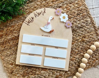 May include: Wooden baby announcement board with a white goose and pink flowers. The board has spaces to fill in the baby's name, date, time, weight, and length. The text "Hello World" is written on the top of the board.
