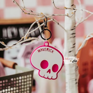 May include: A white and pink skull-shaped keychain with the word "MAVERICK" in white lettering. The keychain is hanging from a black metal clip and ring. The background includes a decorative tree and a pink and white checkered pattern.