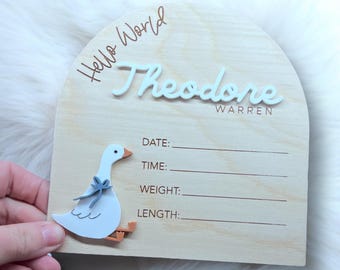 May include: Wooden baby announcement plaque with a white goose cutout, the text "Hello World" in brown lettering, and the name "Theodore Warren" in white lettering. The plaque has spaces to fill in the baby's date, time, weight, and length.