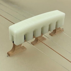 May include: A close-up of a banjo bridge. The bridge is made of light-colored wood and has a white, rectangular top with arched cutouts. The banjo strings are visible, strung across the bridge.