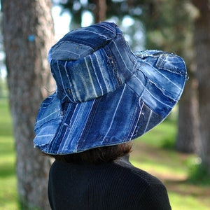 May include: A wide-brimmed hat made from blue denim fabric with a patchwork design. The hat is worn by a person standing in a grassy area with trees in the background.