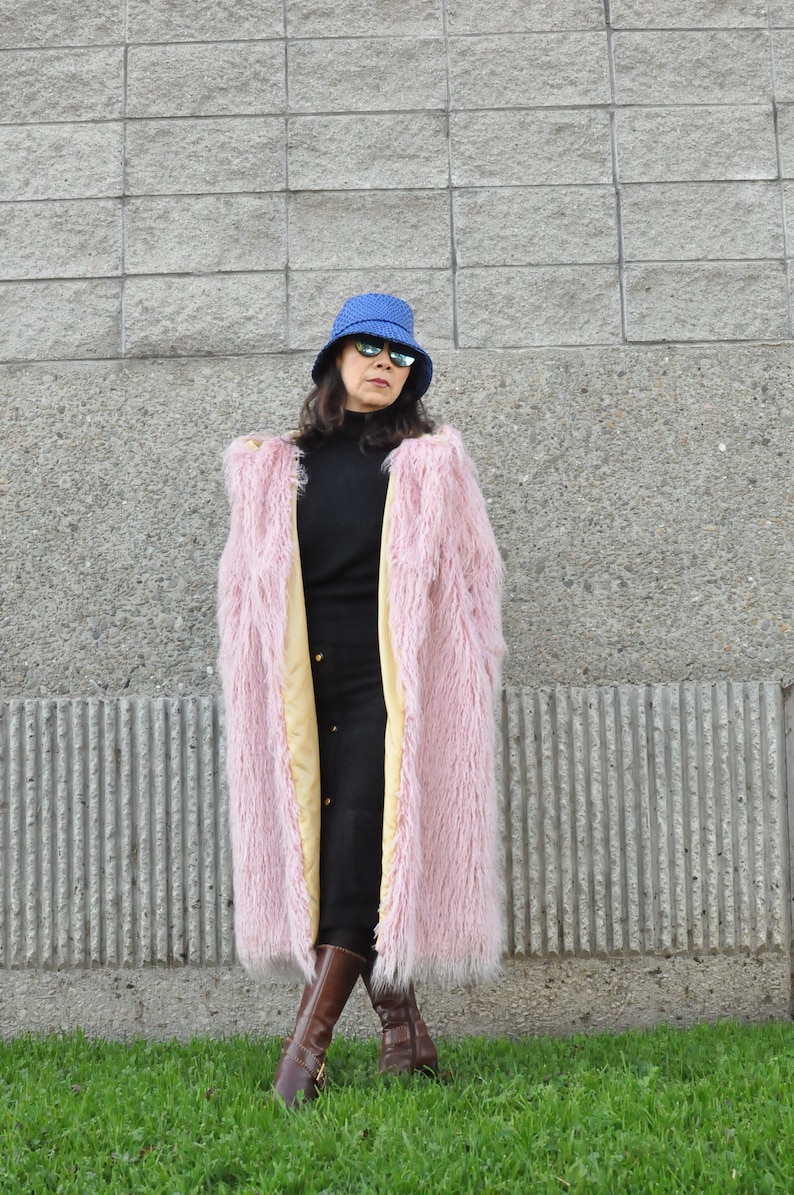 Puede incluir: Una mujer con un abrigo de pelo rosa con forro amarillo, un sombrero de cubo azul y botas marrones. Est&aacute; de pie frente a una pared de hormig&oacute;n gris.