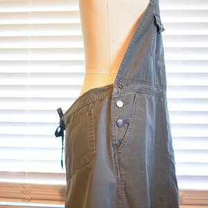May include: A green denim overall dress with a cinched waist and adjustable straps. The dress has a single front pocket and a button closure.