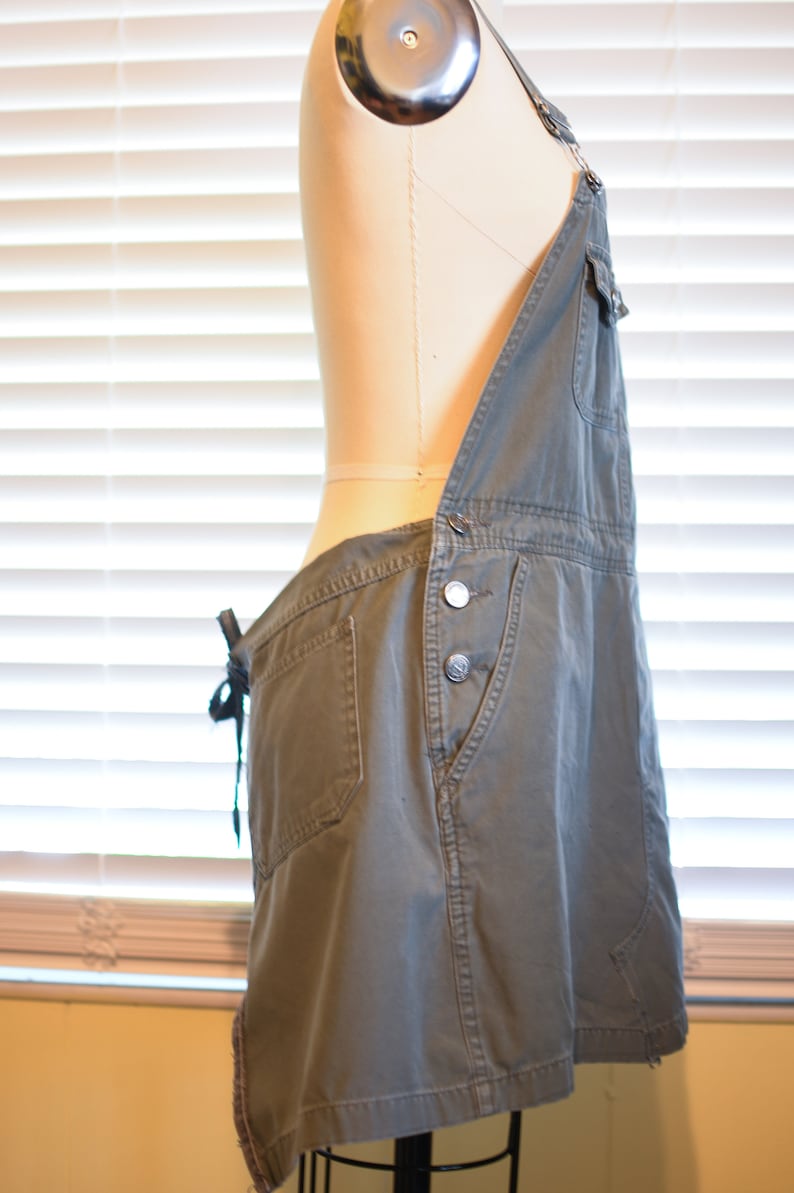May include: A green denim overall dress with a cinched waist and adjustable straps. The dress has a single front pocket and a button closure.