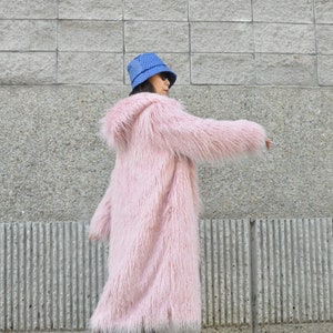 Puede incluir: Una mujer con un abrigo rosa de pelo con capucha, un sombrero azul de pescador y botas marrones. Est&aacute; de pie frente a una pared de ladrillo gris.