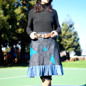 Puede incluir: Una mujer con un jersey negro de cuello alto, una falda vaquera azul con un diseño de patchwork azul y turquesa, un cinturón marrón, un sombrero azul y botas marrones. Está de pie sobre una superficie verde y azul.