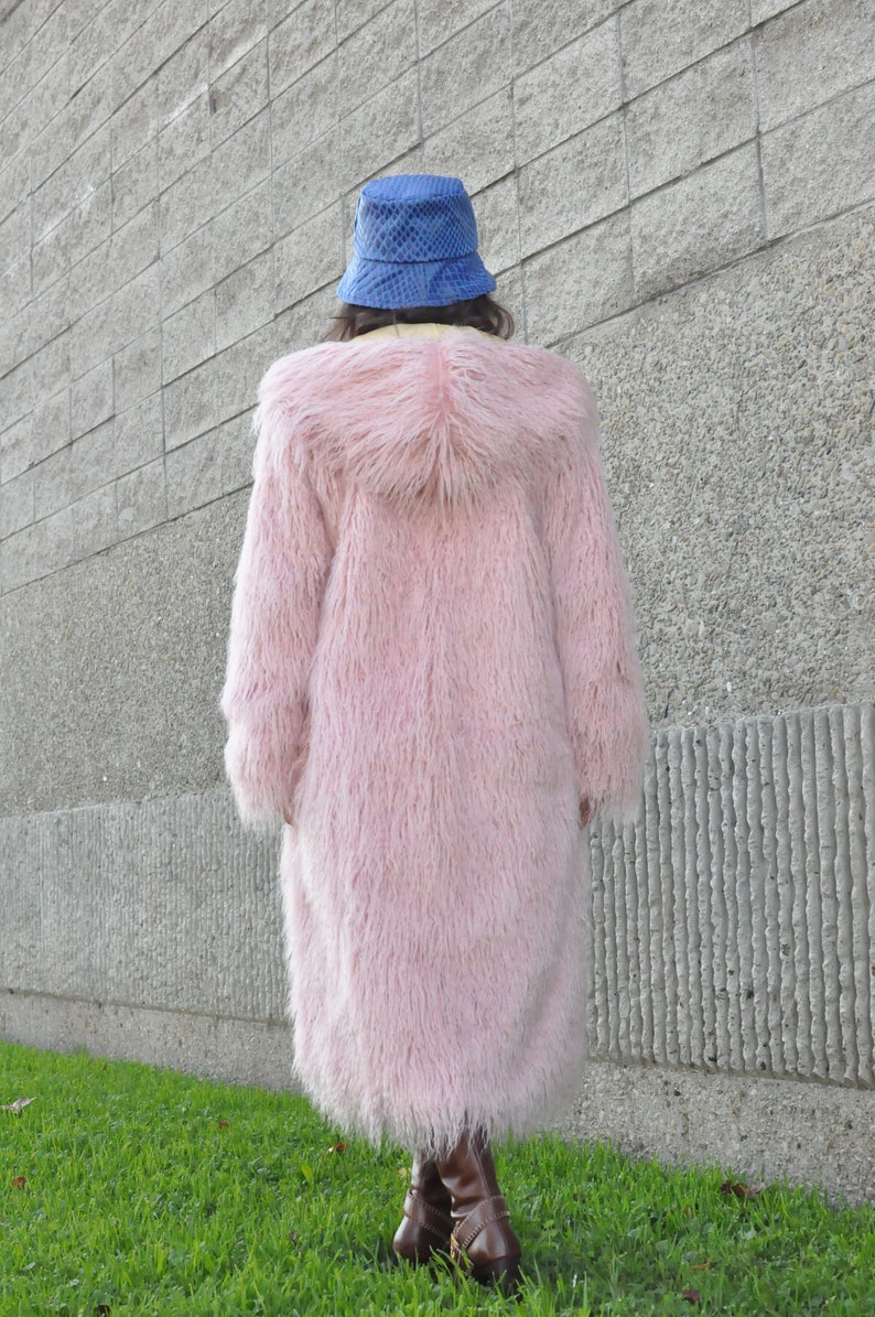 Puede incluir: Una larga y esponjosa chaqueta de piel sint&eacute;tica rosa con capucha. La persona que lleva la chaqueta lleva un sombrero de cubo azul y botas marrones.